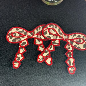 May include: Three leopard print bows with red stitching and a heart design. The bows are attached to a black fabric background.