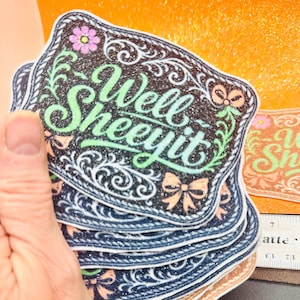 May include: A stack of glittery stickers with a western-style design. The stickers feature the phrase "Well Sheeeyit" in green script, surrounded by floral and bow accents. The background is black with glitter.