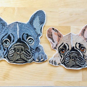 May include: Two embroidered patches in the shape of French Bulldog faces. One is blue and gray, the other is tan and brown. Both have detailed stitching, large eyes, and are lying down.