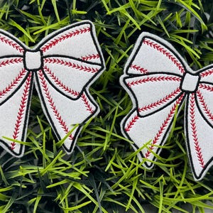 May include: Two white felt bows with red stitching, resembling a baseball, on a green background.