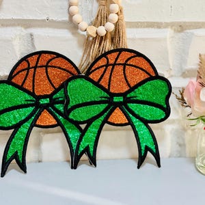 May include: Two decorative basketball ornaments with orange glittered basketballs and bright green bows with black outlines. The ornaments are displayed against a white brick wall, with a vase of flowers to the right.