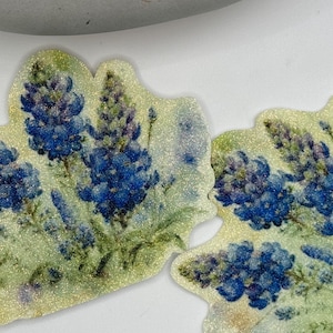 May include: Two leaf-shaped embellishments featuring a watercolor design of bluebonnet flowers. The flowers are a vibrant blue with green foliage, set against a light green and glittery background. The edges are outlined with a shimmering gold.