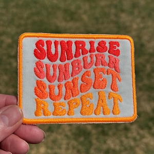 May include: Embroidered patch with the words "Sunrise Sunburn Sunset Repeat" in orange and red lettering. The patch has a white background with an orange border. The patch is rectangular in shape.