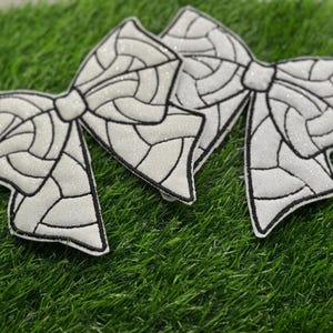 May include: Three white glittery bow appliques with black outlines, arranged on a bed of green artificial grass. The bows have a stained glass design, adding a touch of elegance.