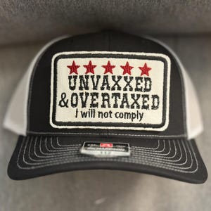 May include: Black and white trucker hat with a rectangular patch featuring the text "UNVAXXED & OVERTAXED I will not comply" in black lettering and five red stars. The hat has a curved brim and a white mesh back.