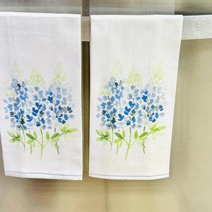 May include: Two white kitchen towels with blue and green watercolor floral designs. The towels feature a cluster of bluebonnet flowers, a Texas state flower.