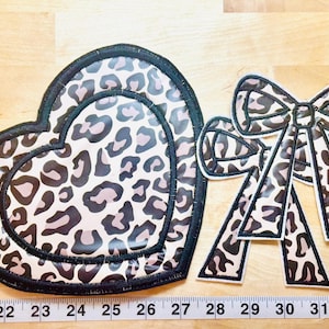 May include: Two leopard print patches: a heart shape and a bow shape. The heart patch is approximately 10 inches wide. Both patches have a black border and a light pink and brown animal print design.