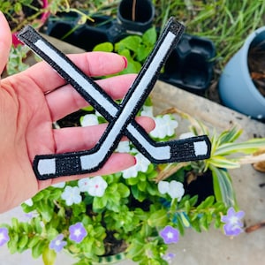 May include: Two black and white hockey stick patches are crossed in the image. The patches have a white stripe down the center and are held in a hand. The background is a garden with green plants and flowers.