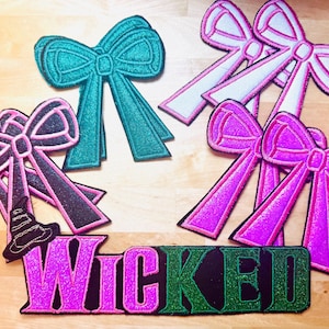 May include: A collection of glittery fabric patches featuring bow shapes in teal, pink, and white, along with a black witch hat and the word "WICKED" in pink and green. These patches are likely for crafting or embellishment.