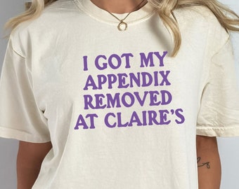 May include: A cream colored t-shirt with the text "I got my appendix removed at Claire's" printed in purple.