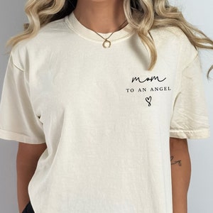 May include: A woman wearing a cream colored t-shirt with the text "mom to an angel" and a heart symbol. She is also wearing black biker shorts.