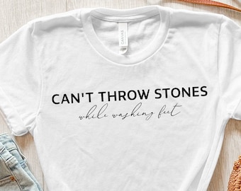 May include: White t-shirt with the text "Can't throw stones while washing feet" in black lettering.