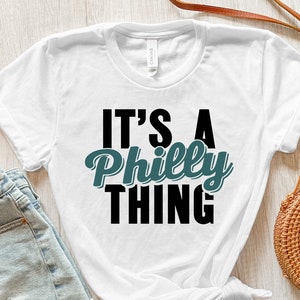 May include: White t-shirt with the text "It's a Philly Thing" in black and teal lettering. The shirt is laid out on a white wooden surface with a pair of light blue denim shorts and a brown wicker bag.