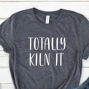 May include: A dark gray t-shirt with the text "TOTALLY KILN IT" in white, paired with blue jeans and white sneakers. The shirt is knotted at the waist, suggesting a casual style.