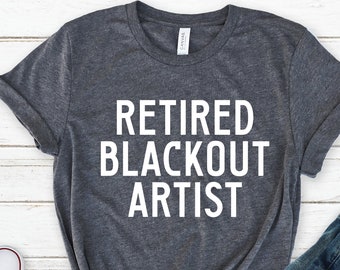 Może przedstawiać: Szary t-shirt z nadrukiem "Retired Blackout Artist" w kolorze białym na przodzie. Koszulka leży płasko na białym drewnianym tle z parą białych butów sportowych i niebieskimi dżinsami.