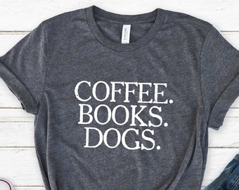 May include: A dark gray t-shirt with the text "COFFEE. BOOKS. DOGS." printed in white on the front. The shirt is laid flat on a white wooden surface with a pair of white sneakers and a pair of blue jeans.