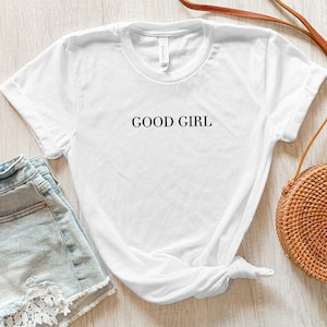May include: A white t-shirt with the text "GOOD GIRL" in black lettering. The shirt is paired with light blue denim shorts with lace detailing and a brown woven crossbody bag.