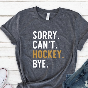 May include: A dark gray t-shirt with the text "SORRY. CAN'T. HOCKEY. BYE." in white letters. The word "HOCKEY" is in gold letters. The shirt is laid out on a white wooden surface with a pair of white sneakers and a pair of blue jeans.