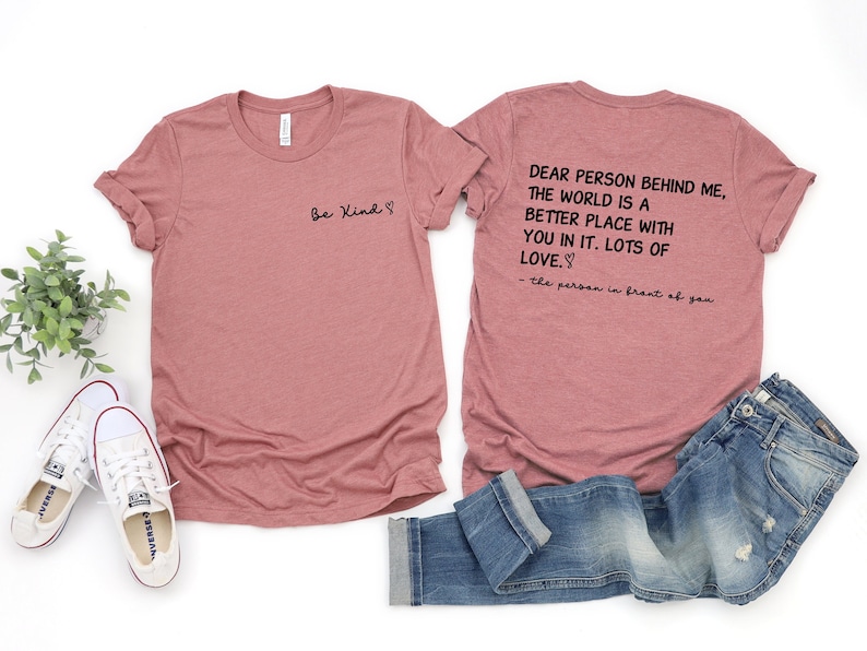 May include: Two pink t-shirts with a message on the back of one shirt that reads "Dear person behind me, the world is a better place with you in it. Lots of love. - the person in front of you". The front of the other shirt reads "Be Kind".