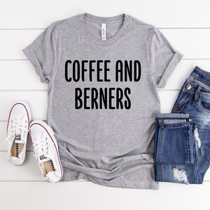 Op de afbeelding: Grijs T-shirt met de tekst "COFFEE AND BERNERS" in zwarte letters. Het shirt wordt gecombineerd met een blauwe jeans en witte sneakers.