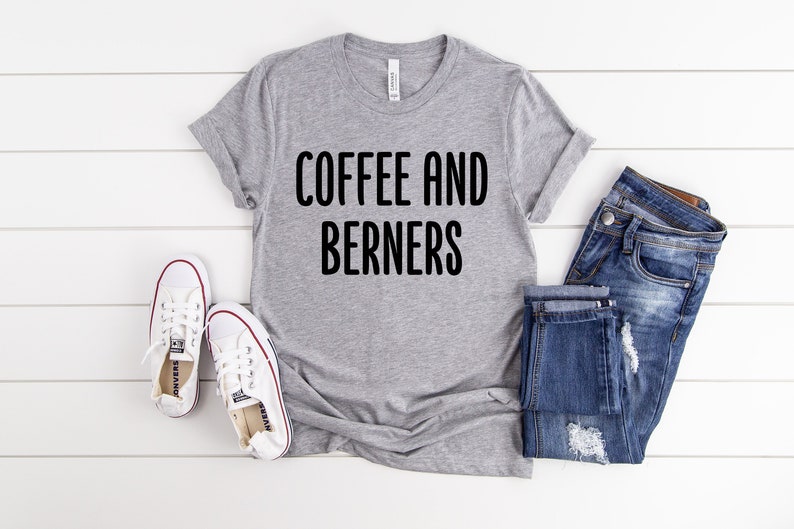 Op de afbeelding: Grijs T-shirt met de tekst "COFFEE AND BERNERS" in zwarte letters. Het shirt wordt gecombineerd met een blauwe jeans en witte sneakers.