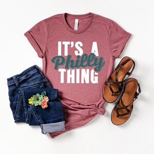 May include: A mauve t-shirt with the text "It's a Philly Thing" in white and teal lettering. The shirt is paired with blue jeans and brown sandals.
