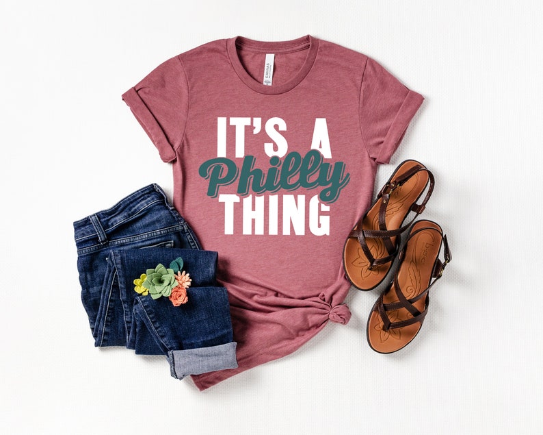 May include: A mauve t-shirt with the text "It's a Philly Thing" in white and teal lettering. The shirt is paired with blue jeans and brown sandals.