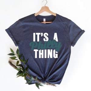 May include: A navy blue t-shirt with white text that reads "It's a Philly Thing". The word "Philly" is in a green font.