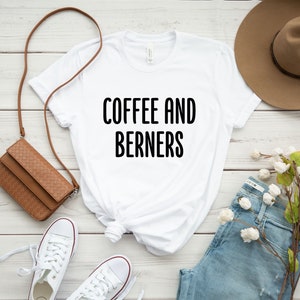 Op de afbeelding: Wit T-shirt met zwarte tekst die "Coffee and Berners" zegt. Het shirt ligt op een wit houten oppervlak met een bruine leren tas, witte sneakers, een blauwe jeans, een bruine hoed en een takje witte bloemen.