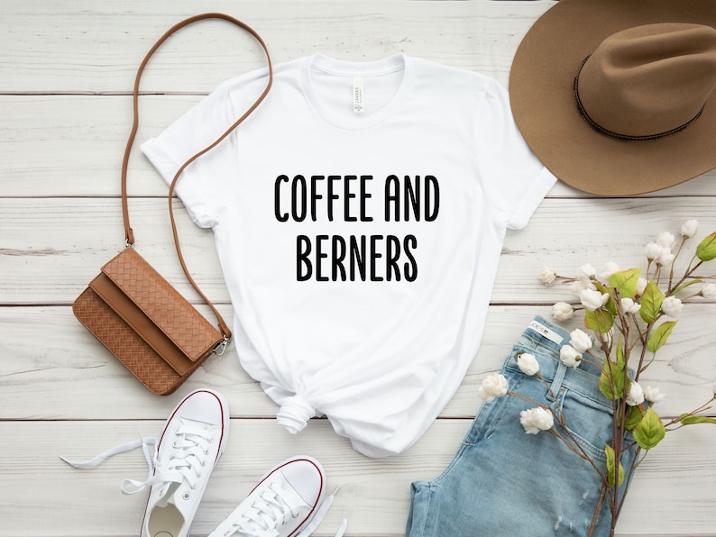Op de afbeelding: Wit T-shirt met zwarte tekst die "Coffee and Berners" zegt. Het shirt ligt op een wit houten oppervlak met een bruine leren tas, witte sneakers, een blauwe jeans, een bruine hoed en een takje witte bloemen.