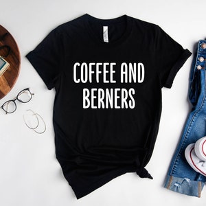 Op de afbeelding: Zwart T-shirt met witte tekst die "Coffee and Berners" zegt. Het shirt ligt plat op een wit oppervlak met een spijkerbroek, een camera en een bril.