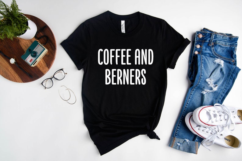 Op de afbeelding: Zwart T-shirt met witte tekst die "Coffee and Berners" zegt. Het shirt ligt plat op een wit oppervlak met een spijkerbroek, een camera en een bril.
