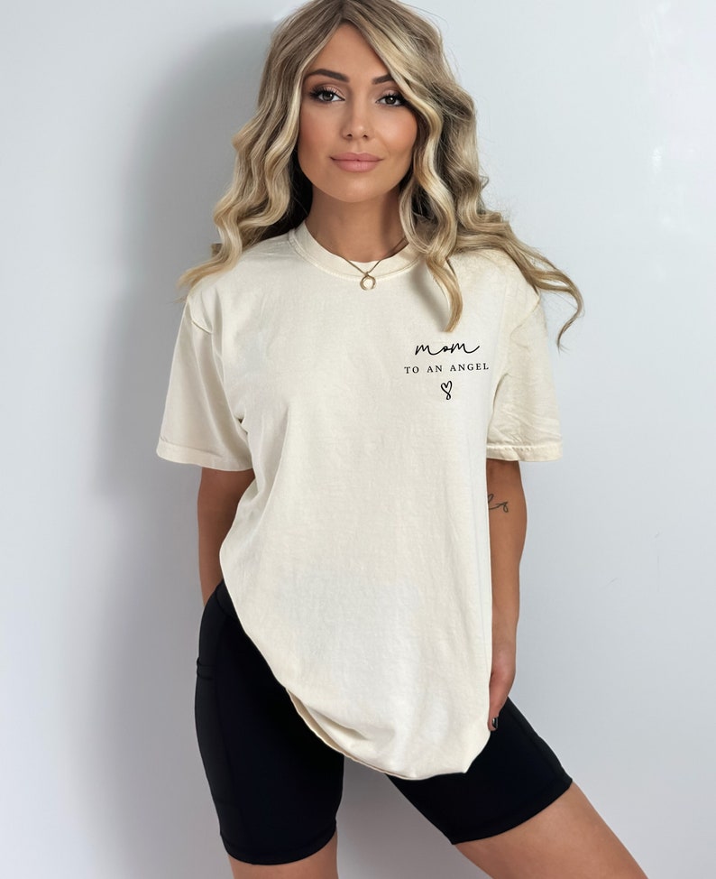 May include: A woman wearing a cream colored t-shirt with the text "mom to an angel" and a heart symbol. She is also wearing black biker shorts.