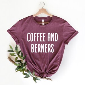 Op de afbeelding: Een maroon t-shirt met de tekst "COFFEE AND BERNERS" in witte letters.