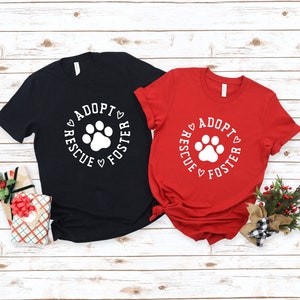 May include: Two t-shirts with a white paw print and the text "Adopt Rescue Foster" in a circle around the paw print. The t-shirts are black and red.
