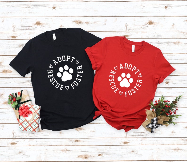 May include: Two t-shirts with a white paw print and the text "Adopt Rescue Foster" in a circle around the paw print. The t-shirts are black and red.