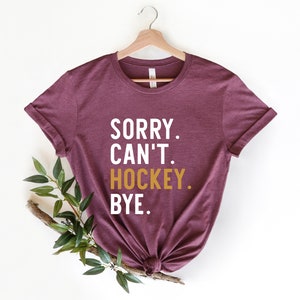 May include: A maroon t-shirt with the text "SORRY. CAN'T. HOCKEY. BYE." in white and gold lettering.