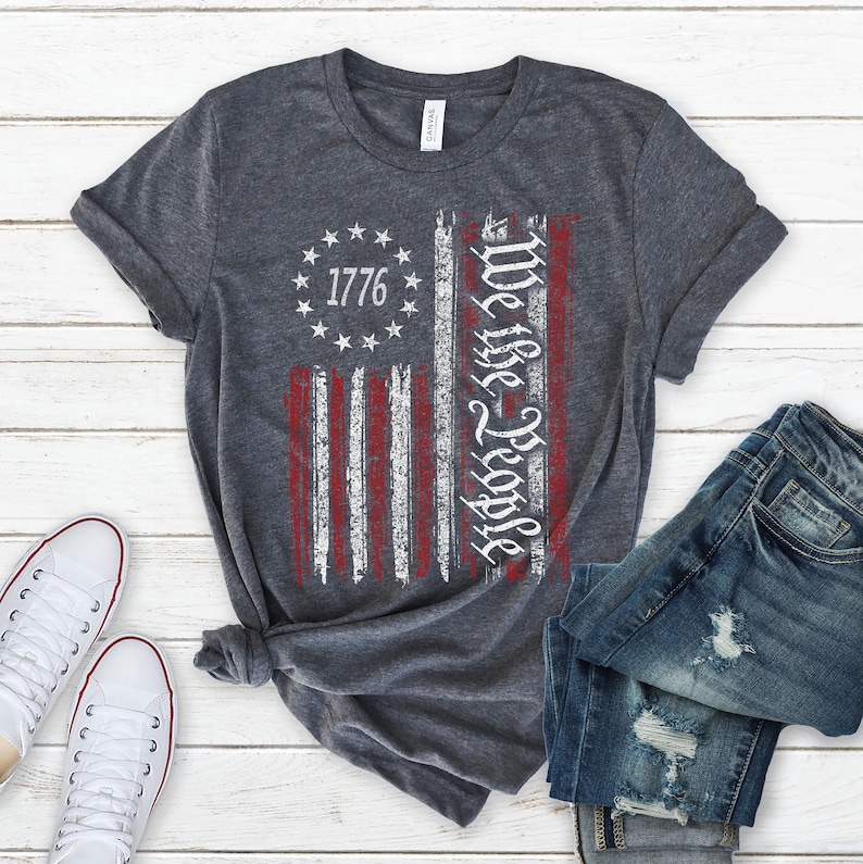Może przedstawiać: Szary t-shirt z nadrukiem postarzanej flagi amerykańskiej. Na fladze widnieje napis "We the People" i rok "1776".