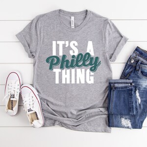 May include: A grey t-shirt with the text "It's a Philly Thing" in white letters with a green and white graphic of the word "Philly".