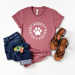 May include: A mauve t-shirt with the text "Rescue Adopt Foster" in a circle with a white paw print in the center. The shirt is paired with blue jeans and brown sandals.