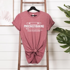 May include: A maroon t-shirt with a white graphic that says "PROCRASTIBAKING" and defines it as a verb: "when you have a million things to do but ignore everything and bake".