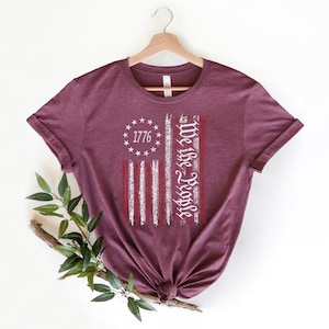 Może przedstawiać: Bordowy t-shirt z nadrukiem przetartej flagi amerykańskiej. Flaga ma białe pasy i czerwone gwiazdy. Tekst "We the People" jest napisany pionowo na fladze. Rok "1776" jest napisany w k&oacute;łku gwiazd.