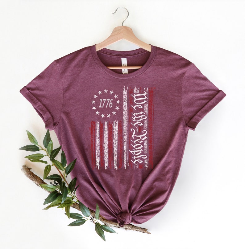 Może przedstawiać: Bordowy t-shirt z nadrukiem przetartej flagi amerykańskiej. Flaga ma białe pasy i czerwone gwiazdy. Tekst "We the People" jest napisany pionowo na fladze. Rok "1776" jest napisany w k&oacute;łku gwiazd.