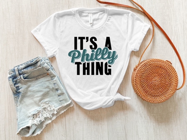 May include: White t-shirt with the text "It's a Philly Thing" in black and teal lettering. The shirt is laid out on a white wooden surface with a pair of light blue denim shorts and a brown wicker bag.