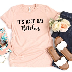 Może przedstawiać: Jasnor&oacute;żowy t-shirt z nadrukiem "It's Race Day Bitches" w kolorze czarnym.