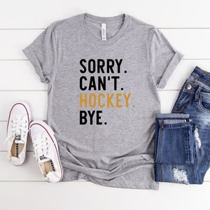 May include: Gray t-shirt with black text that reads "SORRY. CAN'T. HOCKEY. BYE." The word "HOCKEY" is in gold.