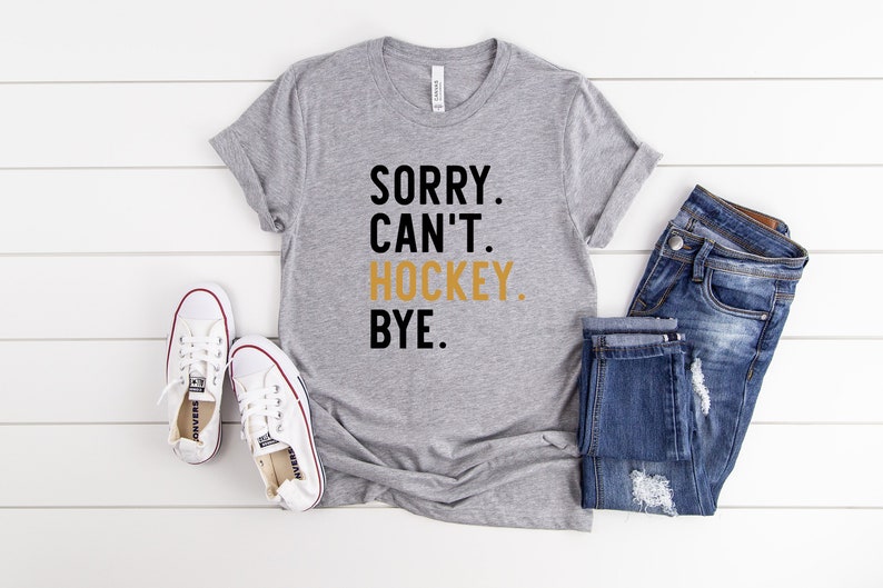 May include: Gray t-shirt with black text that reads "SORRY. CAN'T. HOCKEY. BYE." The word "HOCKEY" is in gold.