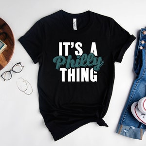 May include: Black t-shirt with white text that reads "It's a Philly Thing" in a green font.