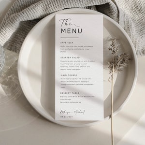 May include: A white wedding menu card with a handwritten script font. The menu includes appetizers, a starter salad, a main course, and a dessert table. The card is on a white plate with a dried flower sprig.