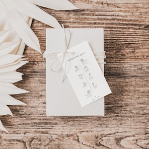 May include: A white gift box tied with twine and a white tag that says "The Wedding Day" with a list of wedding events and times. The box is on a wooden surface with a white palm leaf fan.